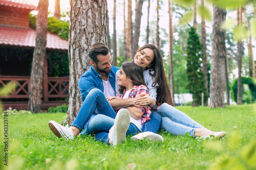 Fototapeta Naklejka Na Ścianę i Meble -  Beautiful happy family while sitting together on the grass and hugging each other, relaxing outdoors on suburban house background.