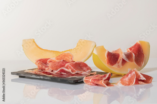 Thin slices of cured ham on a dark plate surrounded by melon slices, one of them with cured ham on top .