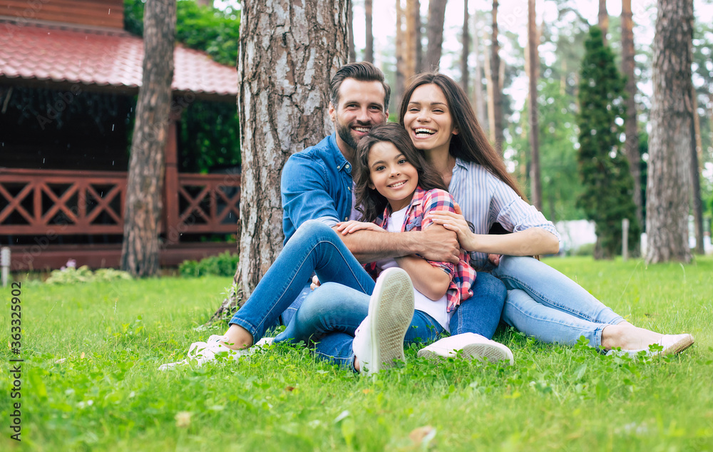 Beautiful happy family while sitting together on the grass and hugging each other, relaxing outdoors on suburban house background. Beautiful happy family while sitting together on the grass and hugging each other, relaxing outdoors on suburban house background.
