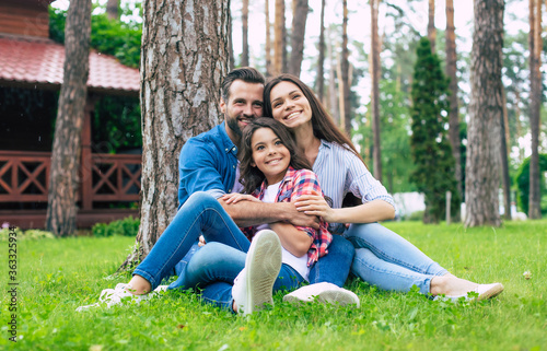 Beautiful happy family while sitting together on the grass and hugging each other, relaxing outdoors on suburban house background.