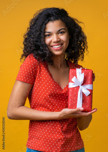 Vertical sshot of happy smiling girl posing with a gift box decorated with satin ribbon in hands isolated on bright colored yellow background photo