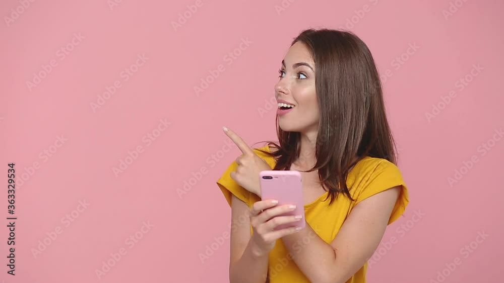 Fun young woman girl 20s in yellow t-shirt posing say wow hold in hand using mobile cell phone pointing fingers aside on workspace isolated on pastel pink background studio. People lifestyle concept Fun young woman girl 20s in yellow t-shirt posing say wow hold in hand using mobile cell phone pointing fingers aside on workspace isolated on pastel pink background studio. People lifestyle concept