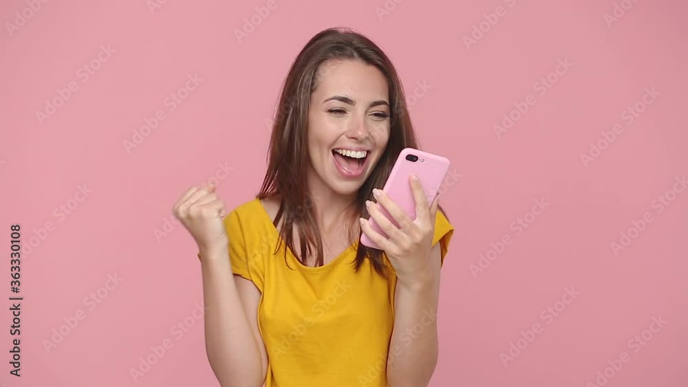 Happy excited young woman girl 20s years old in yellow t-shirt posing hold using mobile cell phone just found out great big win news isolated on pastel pink background studio. People lifestyle concept Happy excited young woman girl 20s years old in yellow t-shirt posing hold using mobile cell phone just found out great big win news isolated on pastel pink background studio. People lifestyle concept