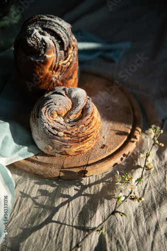 Homemade cruffin breads with dried fruits, cocoa and cinnamon fillings on wooden board and blooming cherry twig on linen tablecloth .