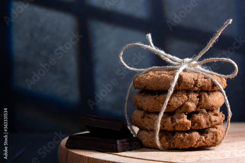 Oatmeal cookies tied with coarse rope with chocolate slices on wooden board. Dark background with incident light from the window frame.
