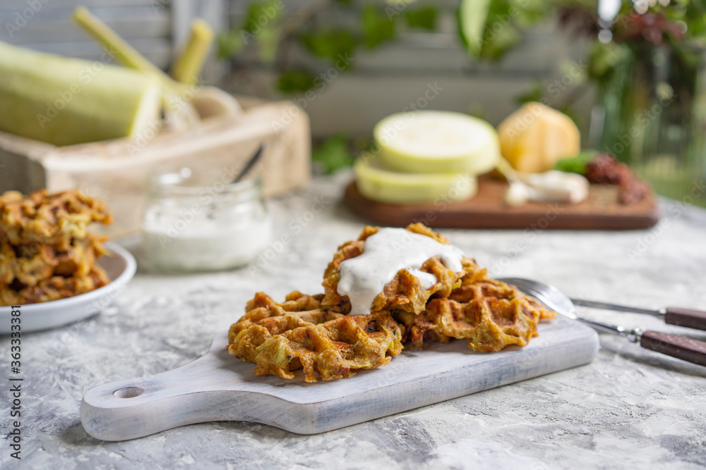 Savory squash or zucchi waffles with cheese, herbs and white sauce. Homemade baked waffles on grey textured table at kitchen, natural light, organic products on background. Selective focus, copy space Savory squash or zucchi waffles with cheese, herbs and white sauce. Homemade baked waffles on grey textured table at kitchen, natural light, organic products on background. Selective focus, copy space