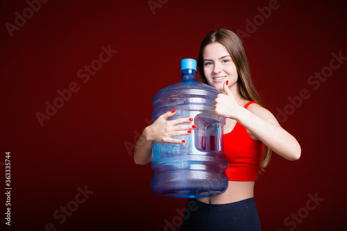 Portrait of a european girl with long hair who hugs a blue gallon of water and shows like on a red background photo