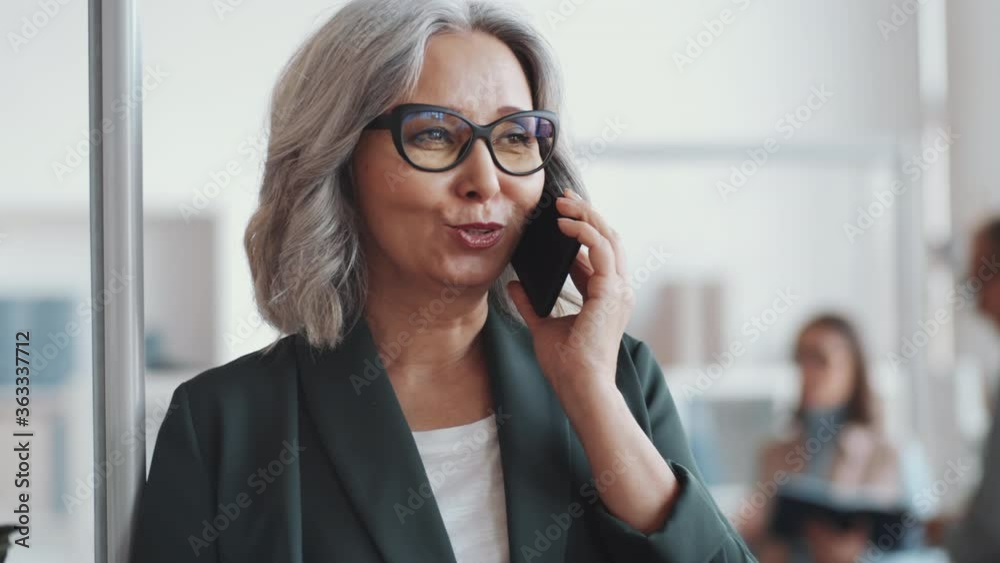 Chest up shot of beautiful senior businesswoman in glasses and formalwear speaking on mobile phone while standing in the office