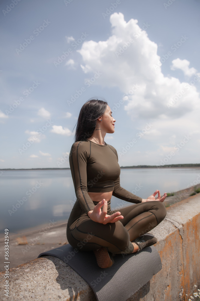 Beautiful brunette in a green suit does yoga by the water. Yoga in the open. Individual yoga class.