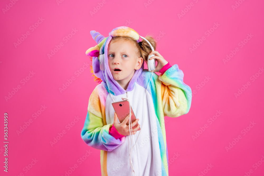 Happy little blonde girl in unicorn kigurumi listens to music holding in hand smatrphone on a pink background