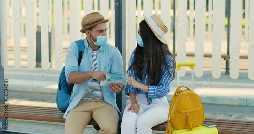 Young Caucasian nice couple of travellers sitting on bench at bus stop and talking while looking for route on map. Summer vacation. Handsome man and beautiful woman in medocal masks with city plan. photo