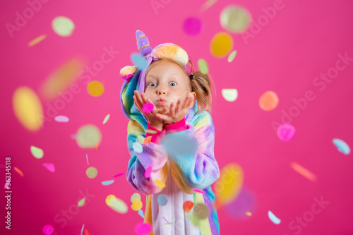 Happy little girl in kigurumi unicorn on a pink background rejoices in multi-colored confetti, space for text