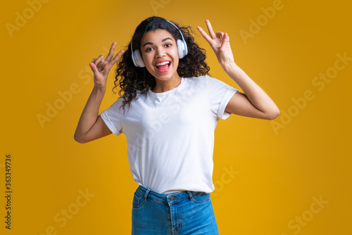 Attractive young dark skinned girl wearing wireless headphones smiling and posing with "v"-gesture