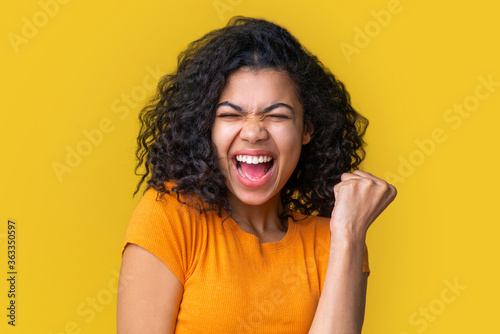 Close up portrait of young euphoric african american woman on bright yellow background showing winner gesture clenching her fist in triumph