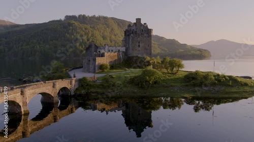 Aerial drone shot tracking around the stunning Eilean Donan Castle in Scotland at sunset, with the lake surrounding it and mountains in the background, and the reflection in the water.