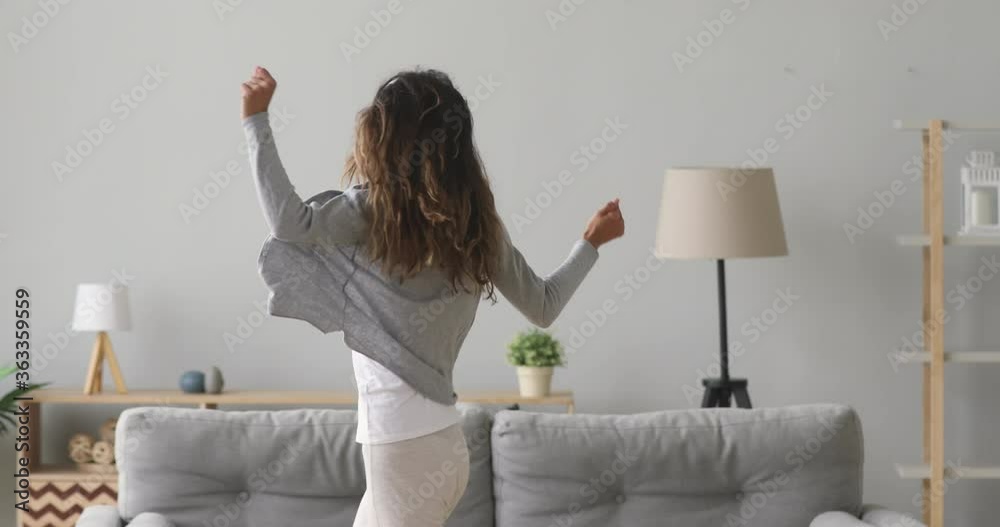 Happy carefree young woman wear wireless headphones dancing alone in modern living room, funny active teen girl jumping moving laughing at home listening to music enjoying new hit song having fun