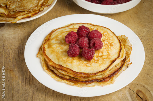 Tasty little thin pancakes with red raspberries on a white plate on a wooden table. Sweet dessert.
