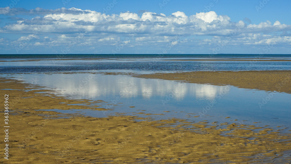 Reflection of clouds and blue sky in the water at low tide with sandy beach in the foreground. Burrum Heads, Queensland, Australia.