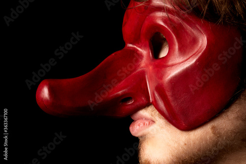 jeune comédien portant un masque de la commedia dell'arte rouge avec un long nez sur fond noir 