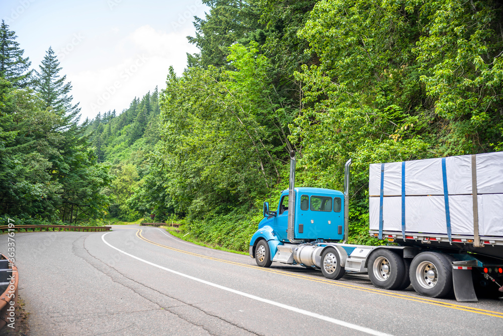 Big rig blue day cab powerful semi truck with high vertical chrome pipes transporting covered lumber wood on flat bed semi trailer running on the winding forest road