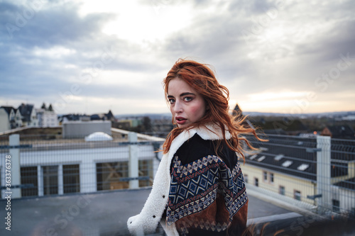 Photo of beautiful girl with long red hairs wearing jacket and looking to camera. Female model standing on flat roof of building. © sebastiancaptures