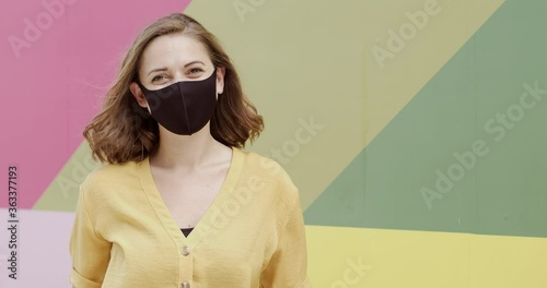 Portrait of pretty female wearing protective face mask looking at camera happy with multi coloured background