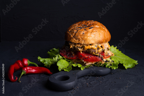 burger with spicy sauce on  black background, large and hearty sprinkled with sesame seeds, red hot pepper lies nearby