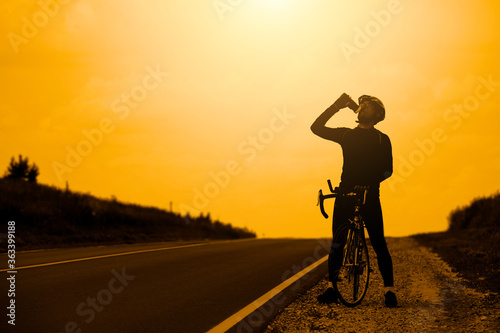 The silhouette of a professional cyclist in sportswear wearing a helmet standing with a Bicycle on an open road against the background of the sunset sky