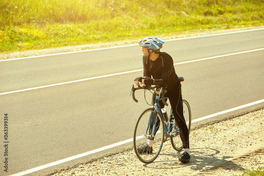 Portrait of a professional cyclist in sportswear wearing a helmet standing with a Bicycle on an open road wearing sunglasses against the sky