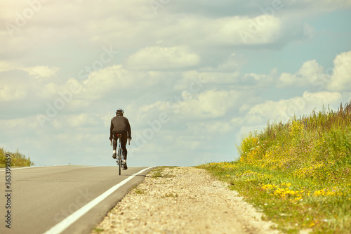 A professional cyclist in sportswear and a helmet rides away on an open road against the sky