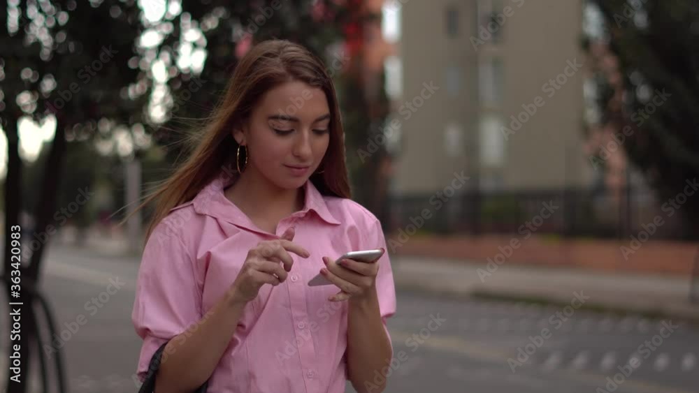 Mujer joven caucásica camina por la calle mientras mira el celular. Revisa las redes sociales, mirando likes y comentarios.