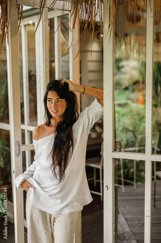 Sensual smiling young woman in white sweater and white pants posing near big white window