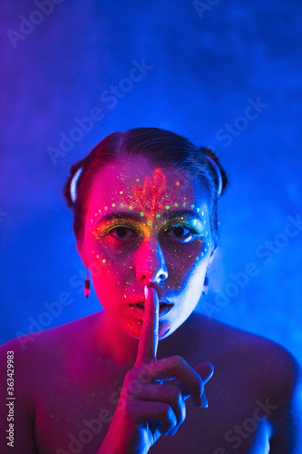 Beautiful woman in with fluorescent makeup shows sign of silence. Glowing paint in neon light.