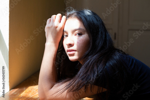 Hispanic young woman lying on her arms at a table with sun rays on her face, young woman thinking at home enjoying the sun, sad and serious woman, looking at the camera.