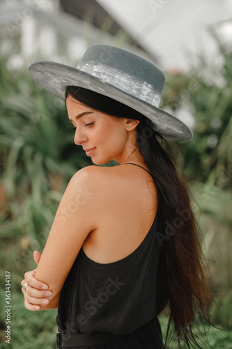 Girl in a dress and a black hat in the field. Young female model wearing a black hat and a black dress. Portrait fashionable girl in elegant hat in trendy black dress look at the camera