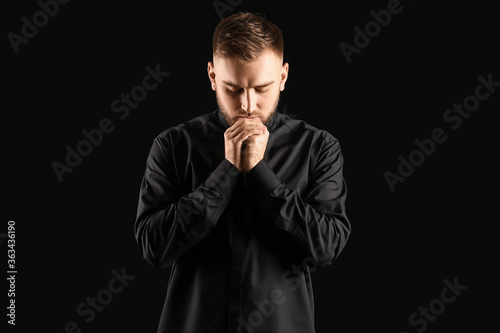 Young priest praying to God on dark background