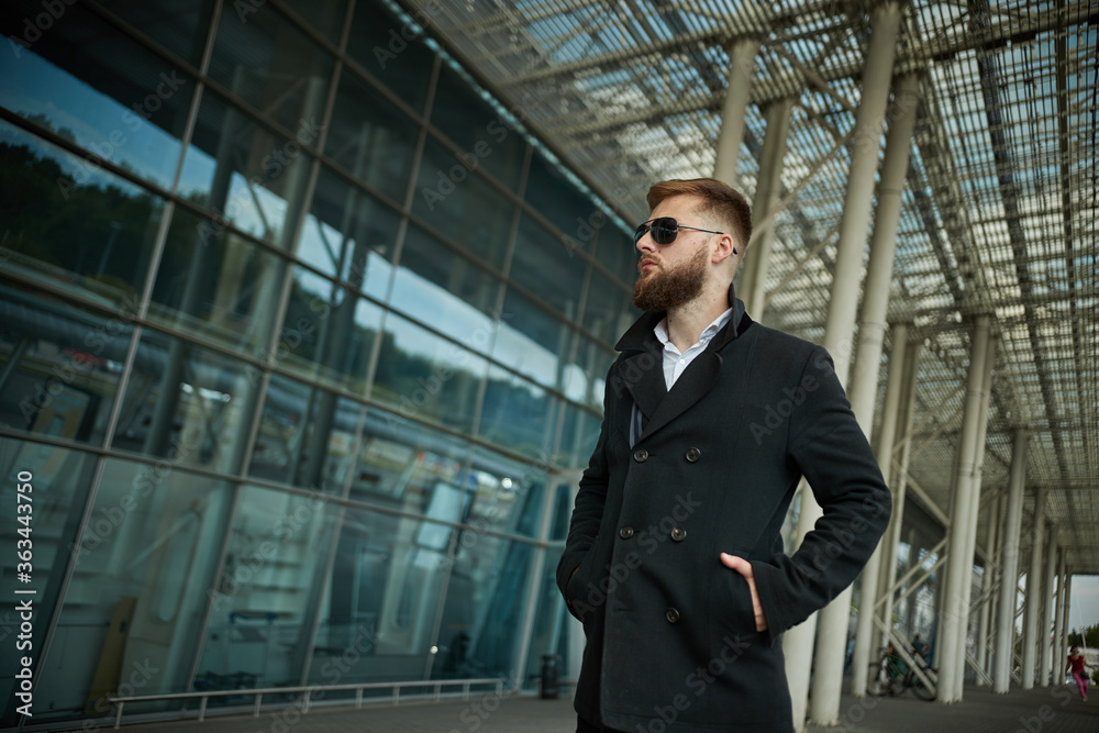 Fototapeta premium Success. Stylish young bearded man in coat, sunglasses businessman standing near the business center, looking far. With hands pockets, pensive and concentrated. Confident look of