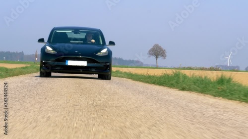 Tesla driving towards camera on a small asphalt road with wind turbines in the background photo