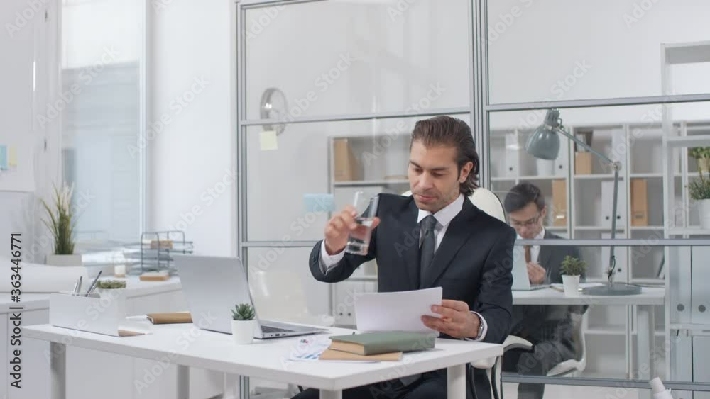 Young adult Caucasian man sitting at office desk drinking water while working with papers, then giving requests to his secretary Young adult Caucasian man sitting at office desk drinking water while working with papers, then giving requests to his secretary