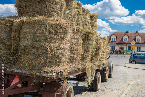 Photo of a tractor hauling hay for animal feed through the cobbled streets of a small town with houses with tiled roofs against a blue sky. The haymaking has begun. Summer. Autumn