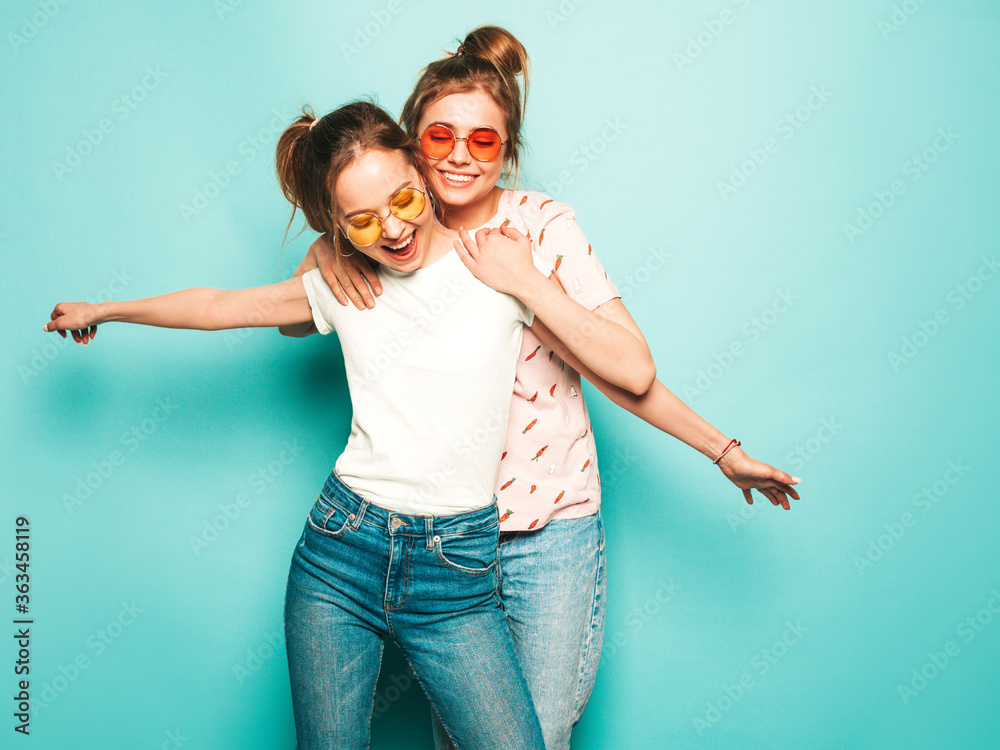 Two young beautiful blond smiling hipster girls in trendy summer hipster jeans clothes. Sexy carefree women posing near blue wall. Trendy and positive models having fun in sunglasses Two young beautiful blond smiling hipster girls in trendy summer hipster jeans clothes. Sexy carefree women posing near blue wall. Trendy and positive models having fun in sunglasses