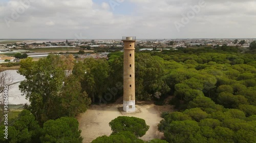 Aerial view of the slightly inland San Jeronimo lighthouse at the mouth of the Guadalquivir River in the Atlantic Ocean in western Analusia. The tower is framed by trees.
