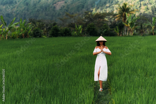 A beautiful girl walks on rice terraces on the island of Bali. A beautiful girl in a white dress and a conical hat walks through green rice fields. Copy space. A woman in a Balinese conical hat