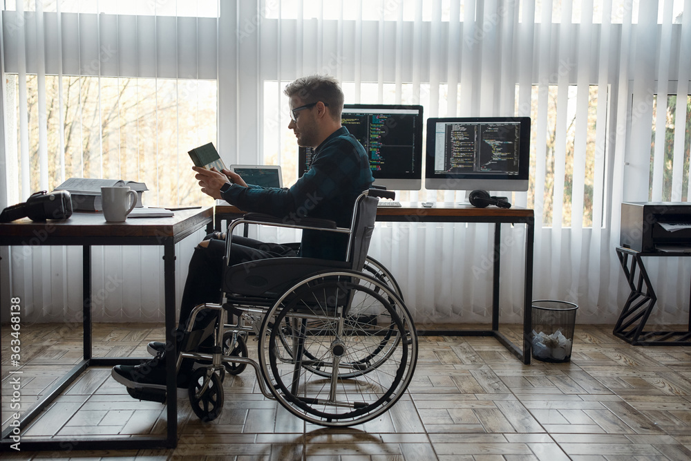 Such an interesting book. Side view of young male web developer in a wheelchair holding a book while sitting at his workplace with multiple computer screens on the background