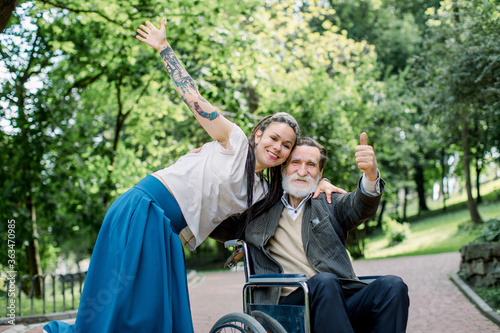 Happy young woman in blue skirt and white shirt, with dreadlocks, smiling, hugging with her senior grandfather on wheelchair in park. Social worker and disabled man on a walk
