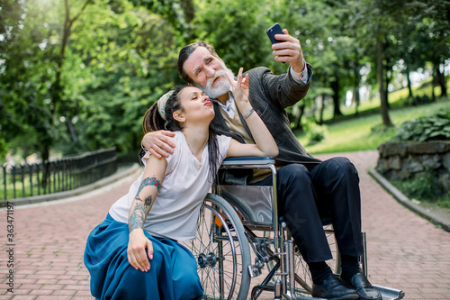 Hipster girl with long dreadlocks and handsome bearded senior man grandfather in wheelchair at park taking selfie. Lifestyle of disabled people. Care giver and patient on a walk