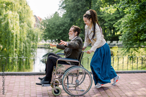 Pretty hipster young woman with dreadlock hair pushing wheelchair with elderly man, having fun in the city park in front of lake. Handicapped man is happy during walk with his granddaughter.