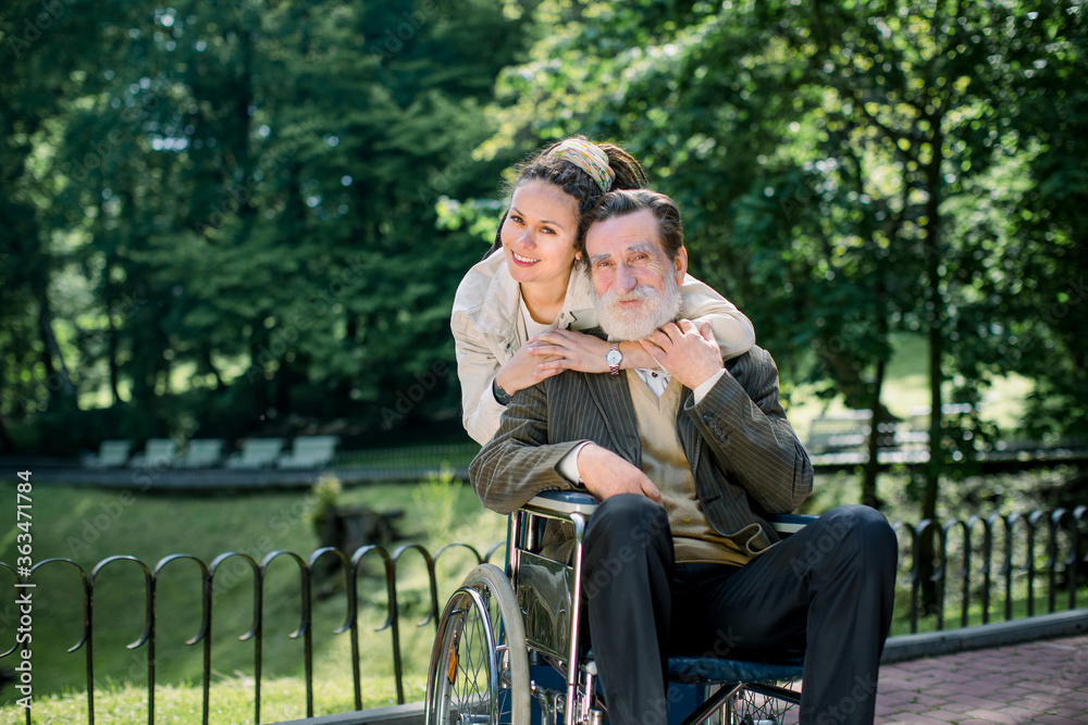 Happy bearded senior man in wheelchair with charming young girl supporting disabled grandparent on walking in park. Grandfather and granddaughter enjoying walk, elderly care concept Happy bearded senior man in wheelchair with charming young girl supporting disabled grandparent on walking in park. Grandfather and granddaughter enjoying walk, elderly care concept