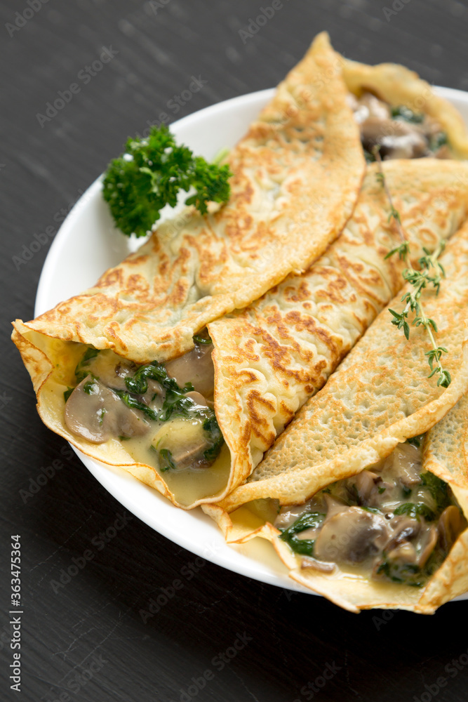 Savory Homemade Mushroom, Spinach and Cheese Crepes on a white plate on a black background, low angle view. Savory Homemade Mushroom, Spinach and Cheese Crepes on a white plate on a black background, low angle view.