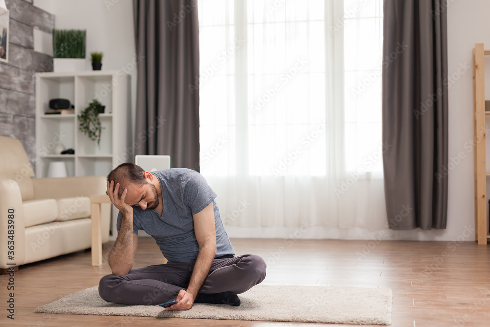 Bored man sitting on carpet in living room browsing on smartphone during quarantine. Bored man sitting on carpet in living room browsing on smartphone during quarantine.